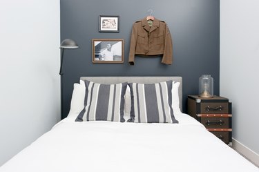 Industrial bedroom with gray accent wall, upholstered headboard, lamp, nightstand, photo, army jacket.
